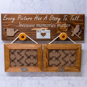 May include: Wooden sign with the text "Every Picture Has A Story To Tell... because memories matter". The sign has a watering can, camera, and bird cutouts. Two brown picture frames hang below the sign.