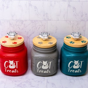 May include: Three ceramic cat treat jars with paw print lids. The jars are red, gray, and teal and each has the words "Cat Treats" written on the front.