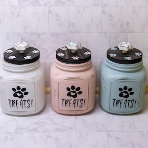 May include: Three glass jars with black lids and paw print designs. Each jar has a white paw print with a heart in the center and the word "Treats!" written below. The jars are painted in white, light pink, and light blue.
