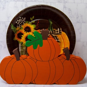 May include: A wooden pumpkin napkin holder with three pumpkins stacked on top of each other. The pumpkins are painted orange with brown stems and green leaves. The holder is decorated with a brown and white patterned paper plate with sunflowers and a pumpkin.