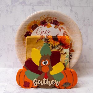 May include: A wooden turkey napkin holder with a brown body, red, green, and yellow feathers, and a pumpkin on each side. The holder has the word "Gather" written on the front. The turkey is holding a paper plate with a fall-themed design and the words "Give Thanks".