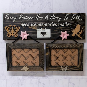 May include: Two dark wood picture frames with a sign above that reads "Every Picture Has A Story To Tell... because memories matter". The sign has a butterfly, camera, and bird cutouts. The frames have a brown patterned background.