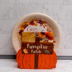 May include: A round paper plate featuring a sunflower and autumn leaf print sits behind a wooden holder shaped like an orange pumpkin. The holder has a brown sign attached that reads "Pumpkin Patch" and holds a stack of napkins with a matching fall design.