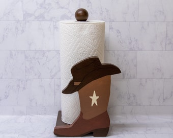 May include: Wooden paper towel holder shaped like a cowboy boot with a brown hat and a white star on the boot. The holder is holding a roll of white paper towels.