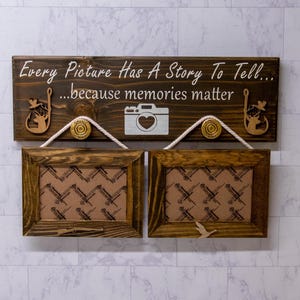 May include: A wooden sign with the text "Every Picture Has A Story To Tell... because memories matter" and two brown picture frames with a patterned background. The sign has a camera and two deer silhouettes.