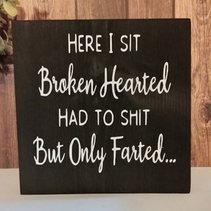 May include: A black wooden sign with white text that reads "Here I sit Broken Hearted Had to Shit But Only Farted..."
