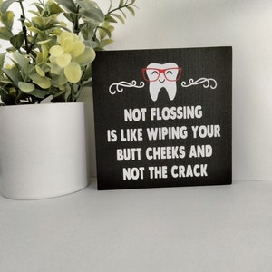 May include: Black chalkboard sign with a white tooth wearing red glasses and the text "Not flossing is like wiping your butt cheeks and not the crack."
