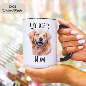 Op de afbeelding: Een witte keramische mok van 444 ml met een zwart handvat en rand. De mok heeft een portret van een lachende golden retriever en de tekst "Goldie's Mom" in het zwart. De linkerbovenhoek heeft een label met "15 oz White/Black".