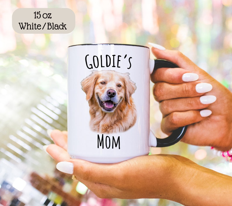 Op de afbeelding: Een witte keramische mok van 444 ml met een zwart handvat en rand. De mok heeft een portret van een lachende golden retriever en de tekst "Goldie's Mom" in het zwart. De linkerbovenhoek heeft een label met "15 oz White/Black".