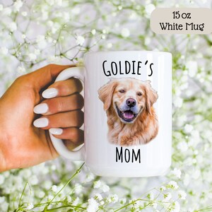 Op de afbeelding: Witte 15 oz mok met een portret van een Golden Retriever en de tekst "GOLDIE'S MOM". De mok heeft een groot handvat. De achtergrond is vervaagd met witte bloemen. De mok wordt door een persoon vastgehouden.