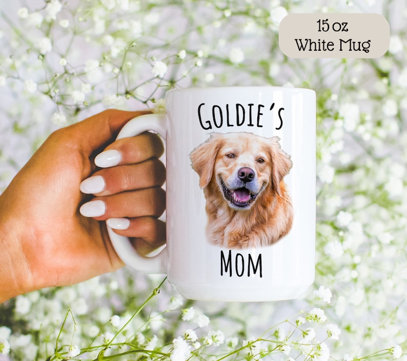 Op de afbeelding: Witte 15 oz mok met een portret van een Golden Retriever en de tekst "GOLDIE'S MOM". De mok heeft een groot handvat. De achtergrond is vervaagd met witte bloemen. De mok wordt door een persoon vastgehouden.