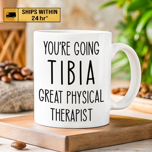 May include: White ceramic coffee mug with black text that reads "You're going to be a great physical therapist." The text "TIBIA" is in a larger font and is centered above the rest of the text.