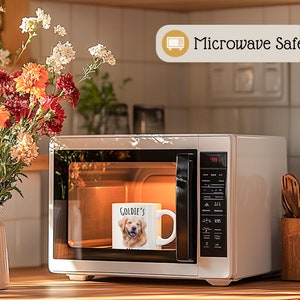Op de afbeelding: Een witte keramische mok met een foto van een golden retriever en de tekst "Goldie's" staat in een magnetron. De magnetron is wit en heeft een digitaal display. De afbeelding bevat ook een vaas met bloemen en de tekst "Microwave Safe".