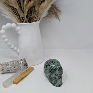 May include: Green and purple gemstone skull carved from a single piece of stone. The skull is sitting on a white surface.
