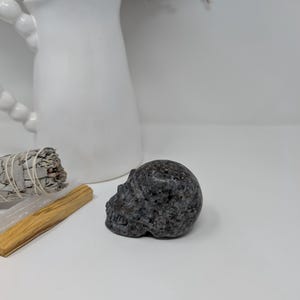 May include: A black and grey speckled stone skull sculpture. The skull is carved in detail and has a smooth, polished finish.