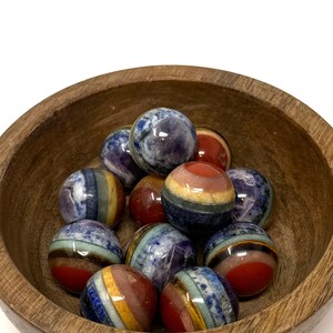 May include: A wooden bowl filled with colorful, layered gemstone spheres. The spheres are made of various stones, including amethyst, lapis lazuli, and tiger's eye.