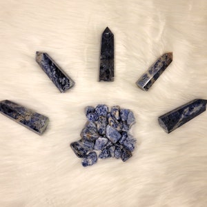 May include: A collection of blue and white sodalite crystals, including tumbled stones and points. The crystals are arranged in a sunburst pattern on a white fluffy surface.