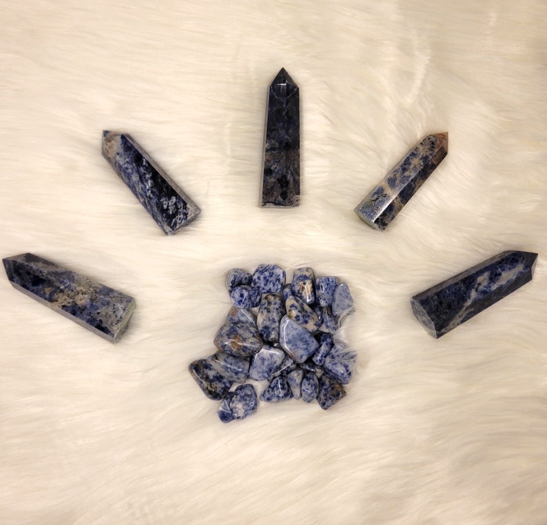 May include: A collection of blue and white sodalite crystals, including tumbled stones and points. The crystals are arranged in a sunburst pattern on a white fluffy surface.