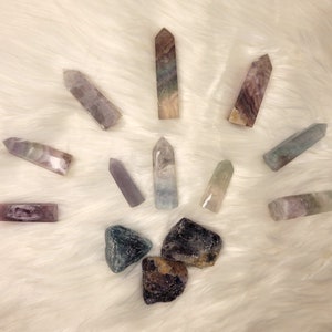 May include: A collection of colorful fluorite crystals, including points and tumbled stones. The crystals are arranged in a starburst pattern on a white fluffy surface.