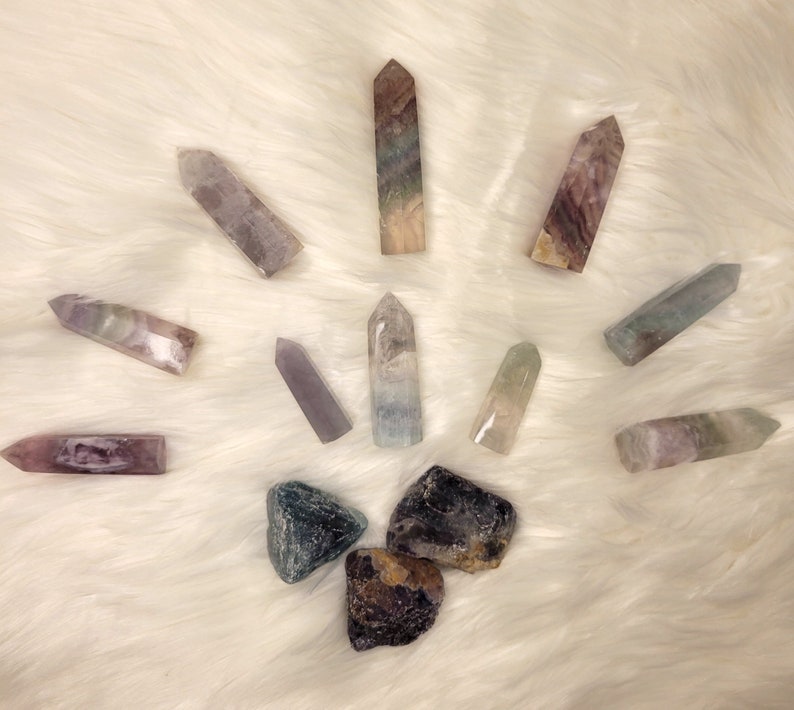 May include: A collection of colorful fluorite crystals, including points and tumbled stones. The crystals are arranged in a starburst pattern on a white fluffy surface.