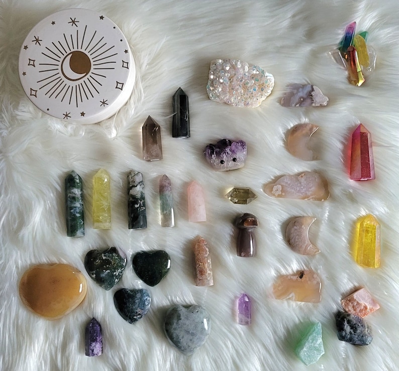 May include: A collection of colorful crystals and stones, including amethyst, rose quartz, clear quartz, and agate. The crystals are arranged on a white fluffy surface.