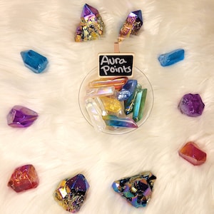 May include: A collection of iridescent aura quartz points in various colors, including purple, blue, green, and yellow. The points are arranged in a bowl and on a white fluffy surface. A small chalkboard sign reads "Aura Points".