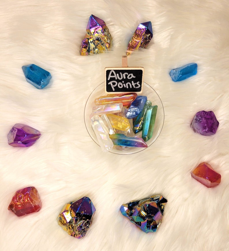 May include: A collection of iridescent aura quartz points in various colors, including purple, blue, green, and yellow. The points are arranged in a bowl and on a white fluffy surface. A small chalkboard sign reads "Aura Points".