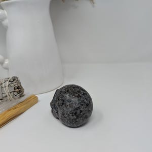 May include: A small, dark grey, polished stone skull. The skull is smooth and has a slightly rounded shape.