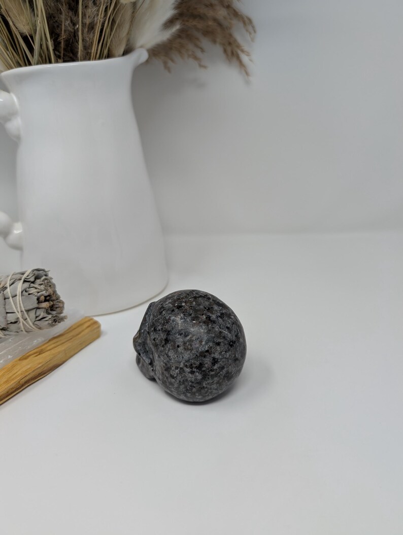 May include: A small, dark grey, polished stone skull. The skull is smooth and has a slightly rounded shape.
