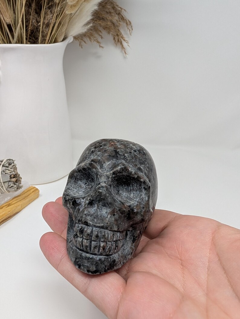 May include: A black and grey granite skull sculpture with detailed features. The skull is carved with a realistic smile and eye sockets.