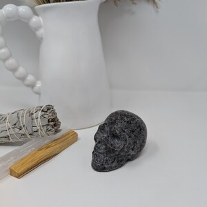 May include: A small, black and grey, carved stone skull. The skull is sitting on a white surface.
