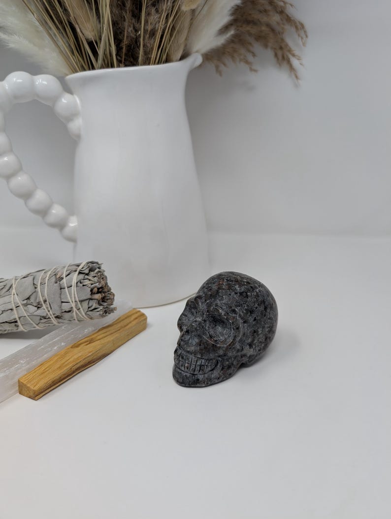 May include: A small, black and grey, carved stone skull. The skull is sitting on a white surface.