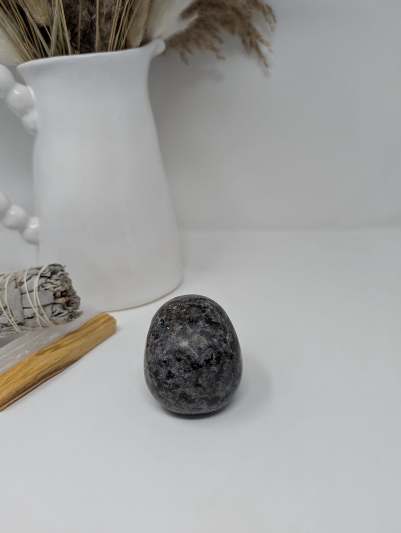 May include: A smooth, dark grey, oval-shaped stone with a speckled pattern. The stone is sitting on a white surface.