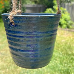 May include: A hanging ceramic planter with a glossy, deep blue glaze and horizontal ribbed texture. The pot features two circular holes near the rim, through which thick, natural-colored jute twine is threaded to allow for suspension.
