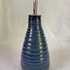 May include: A conical ceramic oil cruet featuring a deep blue glaze with horizontal ribbed textures. The vessel is topped with a stainless steel pourer spout that includes a small black plastic cap, providing a functional design for liquid dispensing.