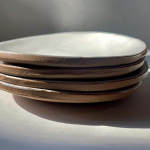 May include: A stack of four small, shallow ceramic plates with an irregular, organic shape. Each plate features a smooth, matte white interior surface and a contrasting, textured, raw brown clay exterior edge that remains unglazed.