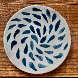 May include: A round ceramic plate with a white base and a hand-painted pattern of dark blue teardrop shapes arranged in a swirling, radial design that radiates from the center toward the outer edge of the plate's circular rim.