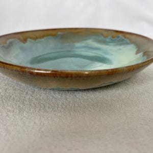 May include: A shallow ceramic bowl featuring a reactive glaze with a blend of earthy brown tones along the outer rim and a soft, muted teal and light blue center. The surface has a smooth, glossy finish with subtle color variations throughout.