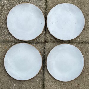 May include: Four white ceramic plates with a brown rim. The plates are arranged in a square pattern on a gray concrete surface.