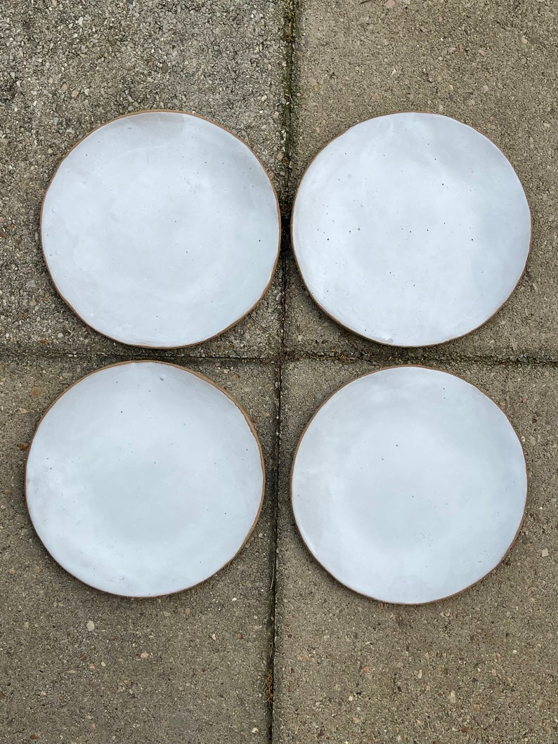 May include: Four white ceramic plates with a brown rim. The plates are arranged in a square pattern on a gray concrete surface.