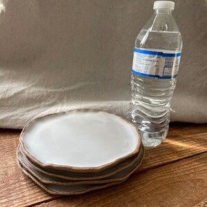 May include: Four white ceramic plates with a brown rim stacked on top of each other. The plates have a slightly wavy edge. A clear plastic water bottle is behind the plates.