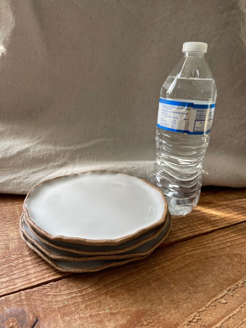 May include: Four white ceramic plates with a brown rim stacked on top of each other. The plates have a slightly wavy edge. A clear plastic water bottle is behind the plates.