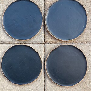 May include: Four black ceramic plates with a slightly raised rim. The plates are arranged in a 2x2 grid on a light gray concrete surface.