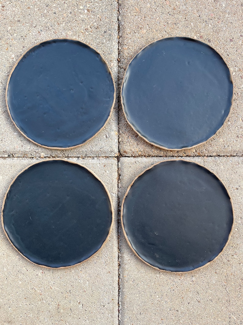 May include: Four black ceramic plates with a slightly raised rim. The plates are arranged in a 2x2 grid on a light gray concrete surface.