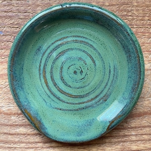 May include: A handmade, round ceramic spoon rest. The spoon rest is glazed in shades of green and blue, with a spiral design in the center. The spoon rest is approximately 6 inches in diameter.
