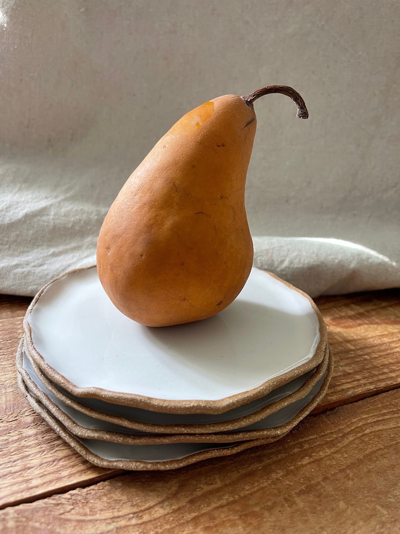 May include: A stack of four white ceramic plates with a brown rim. The plates have a wavy edge and are stacked on a wooden surface. A yellow pear sits on top of the plates.