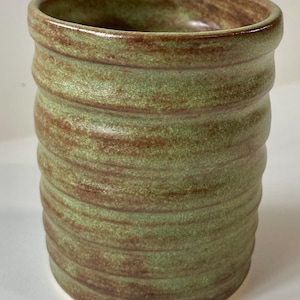 May include: A cylindrical ceramic tumbler featuring a textured, ribbed exterior. The surface displays a mottled glaze with blended shades of sage green and earthy brown, creating a variegated appearance across the entire body of the vessel.