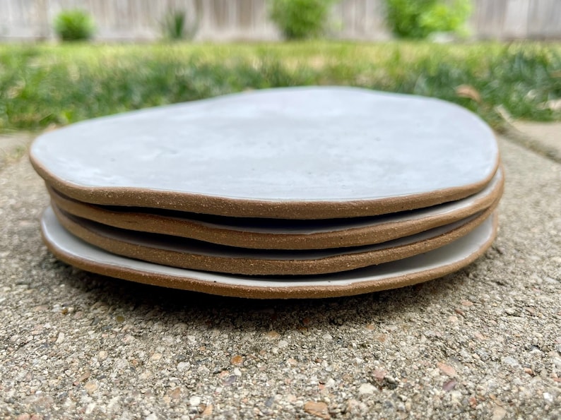 May include: A stack of four white ceramic plates with a brown rim. The plates are slightly irregular in shape and have a matte finish.