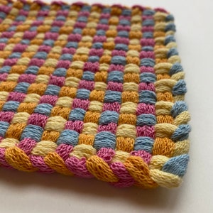 May include: A close-up of a woven textile with a repeating pattern of squares in shades of pink, yellow, blue, and gold. The edges are finished with a braided border in coordinating colors. The textile appears to be handmade.