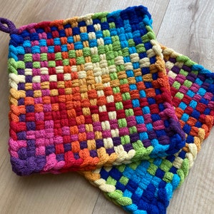 May include: Two colorful woven potholders in a rainbow of hues, including red, orange, yellow, green, and blue. Each square potholder has a loop for hanging. The potholders are made of woven fabric.